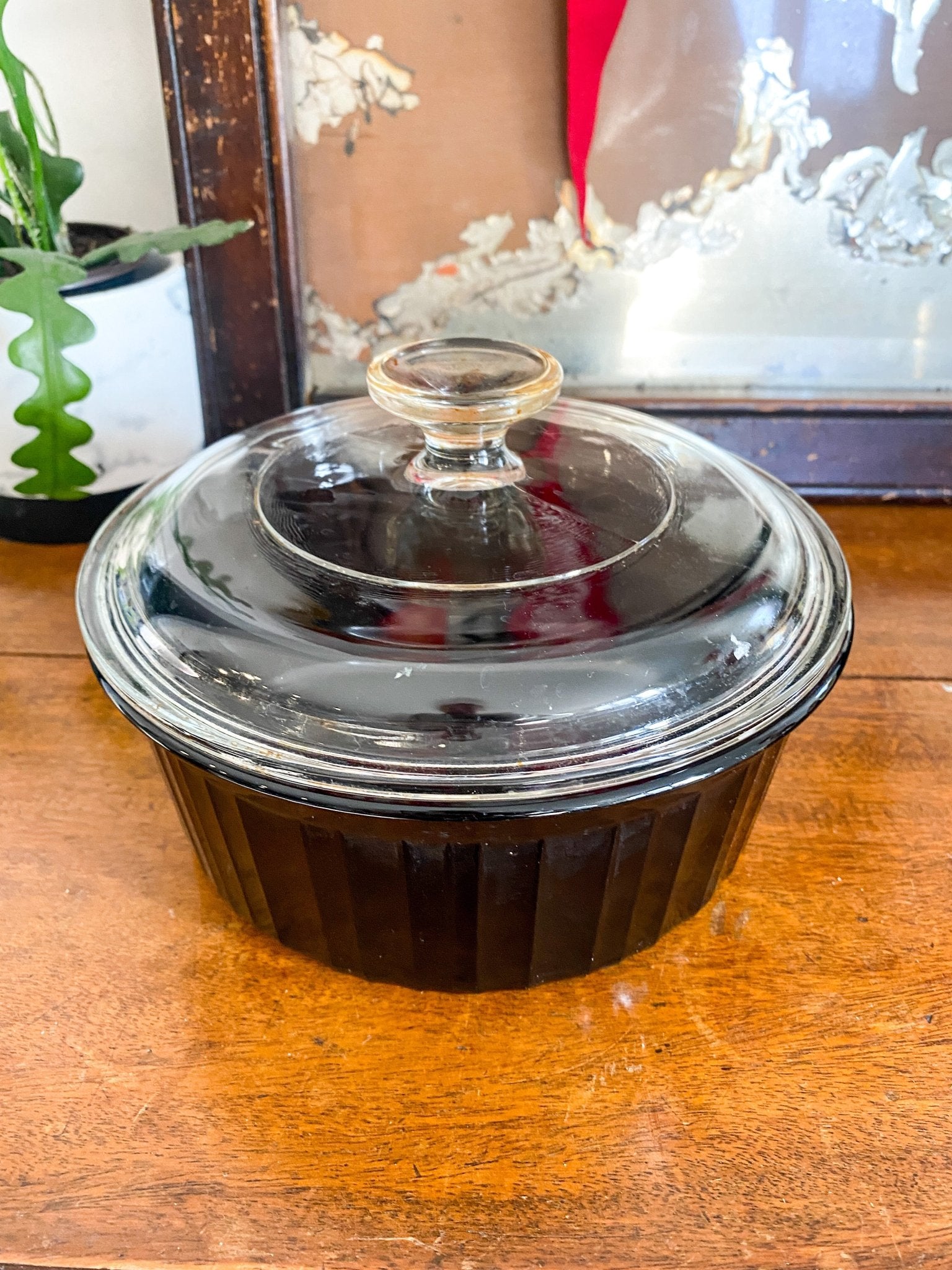 The black dish is shown on an angle from the top to display both the fluted black glass sides and the round, clear top lid with the round handle. It sits on a wooden surface with a plant to the left. 