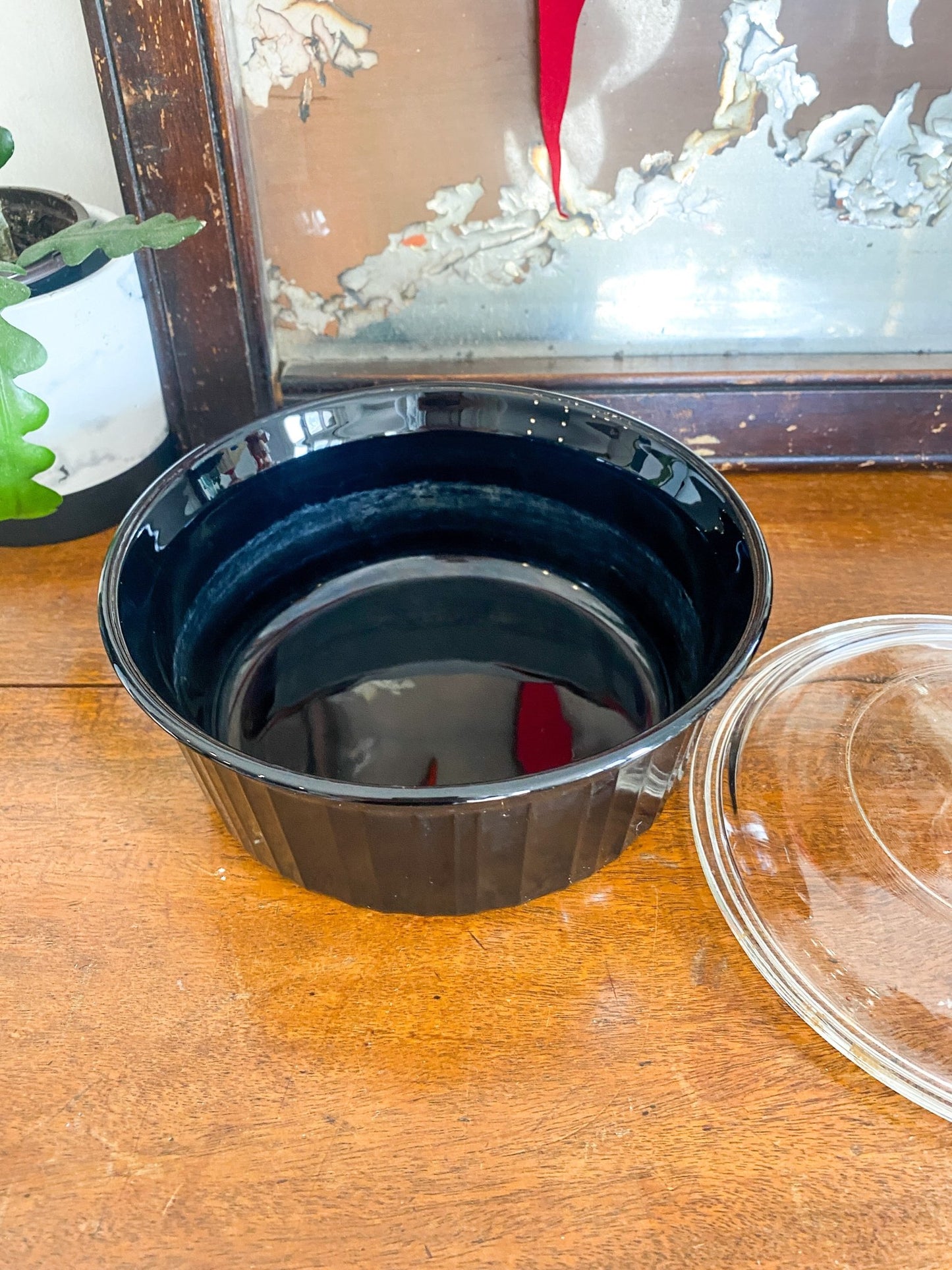 The lid for the dish is off to the right, and the black casserole glass dish is seen front and center. It sits on a wooden table with a plant to the left and a vintage mirror behind. 