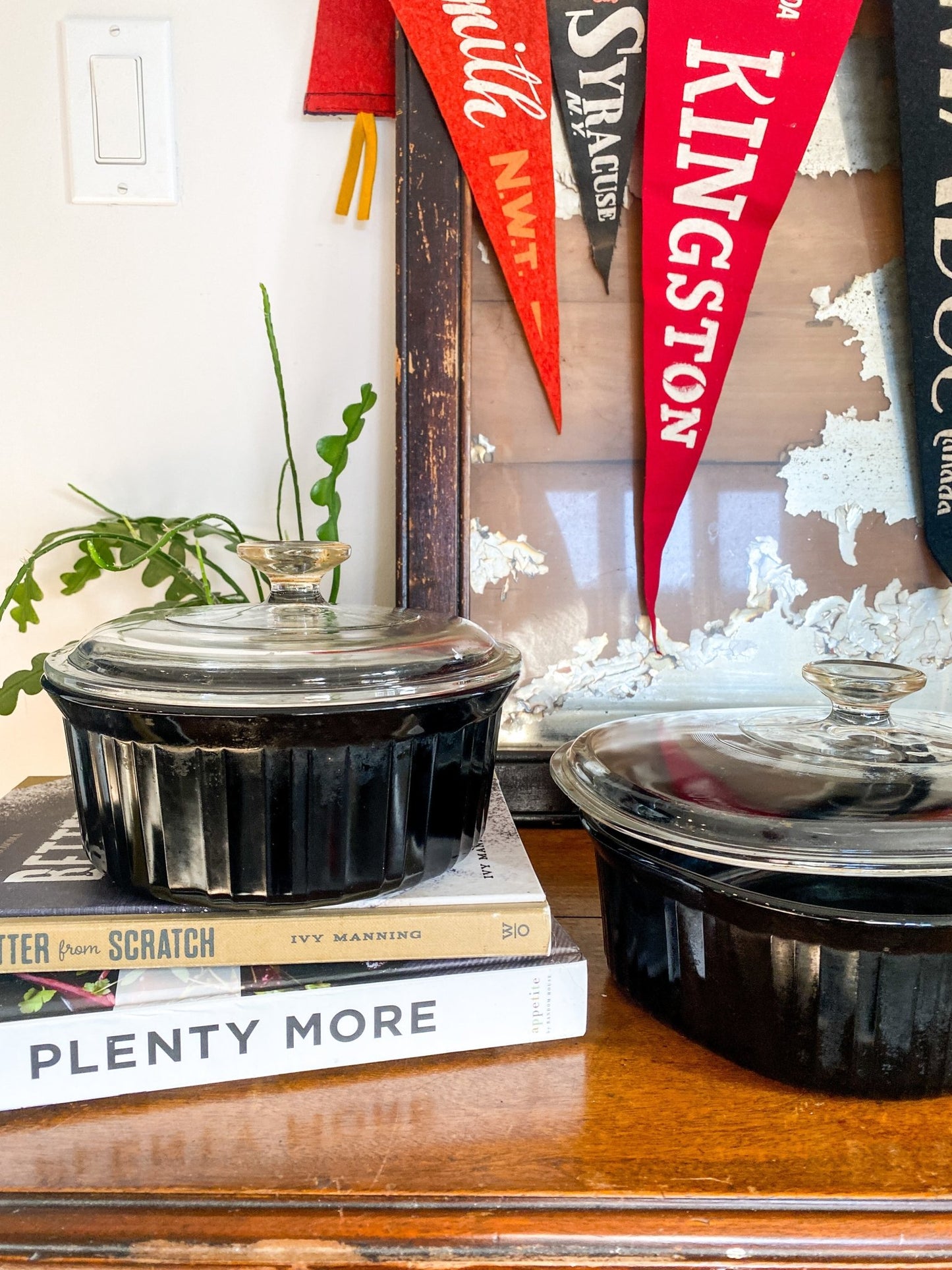 Two black glass casserole dishes are seen here with the lid. The smaller one is on top of 2 cookbooks, Better from Scratch and Plenty More. The larger obtuse one is sitting on a wooden table. There are vintage pennant flags visible in the background