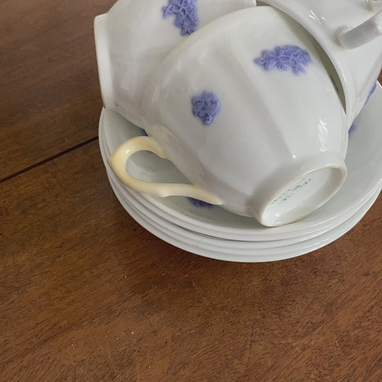 There are three teacups stacked on the saucers within eachother on a wooden table. The teacups are all upright but the handle is facing separate directions. The teacups are on the saucers and show the blue or violet colours. The woman pulls one teacup out to show it off. 