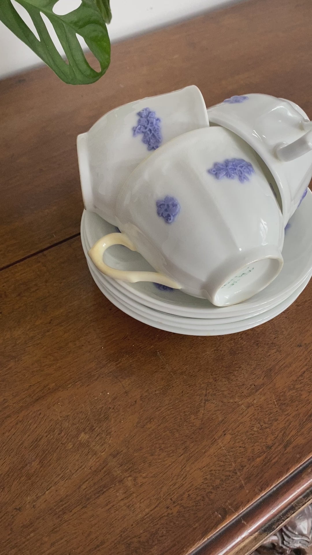 There are three teacups stacked on the saucers within eachother on a wooden table. The teacups are all upright but the handle is facing separate directions. The teacups are on the saucers and show the blue or violet colours. The woman pulls one teacup out to show it off. 