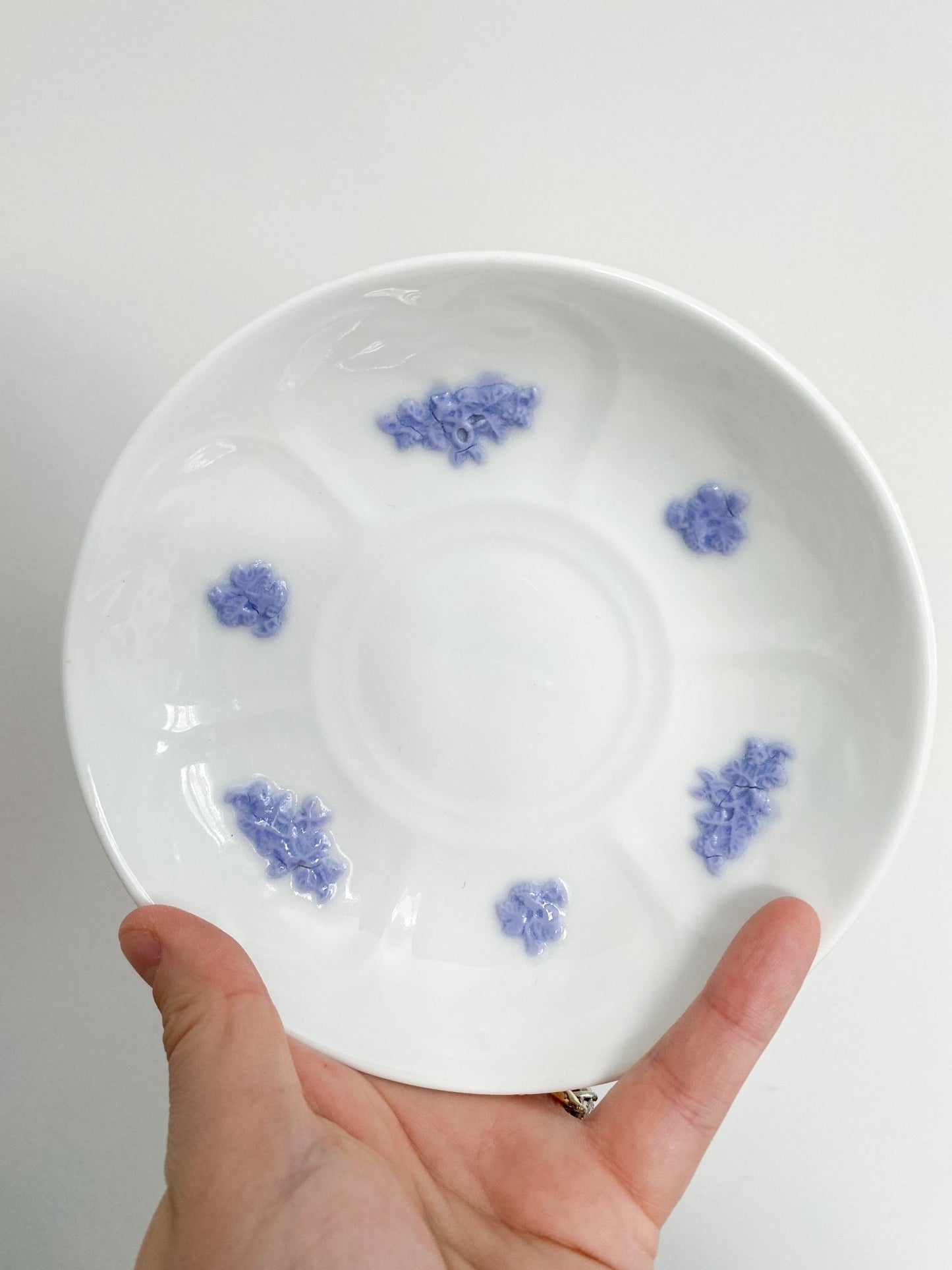 A hand holds the saucer in full view against a white backdrop. The saucer shows the violet flower details, 6 total with 3 smaller flowers and 3 larger flowers.