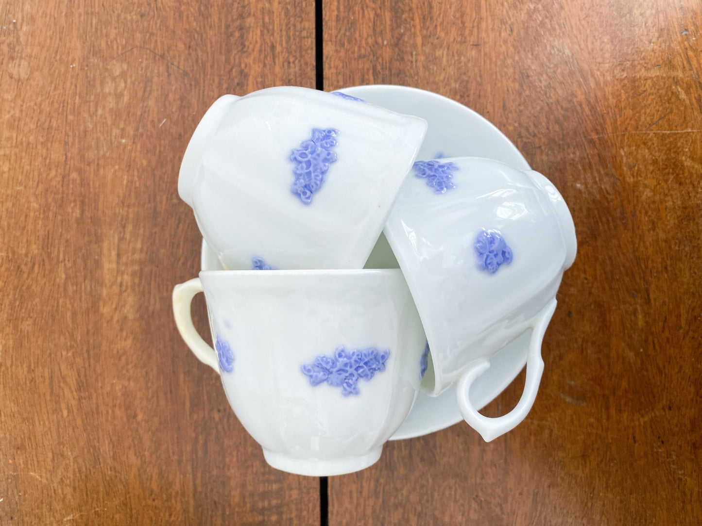Three small tea cups sit on top of saucers in a pattern. They are facing on their sides with the handles in various directions to show off the violet flowers. The china is a white stark contrast in variation to the wooden tabletop. 