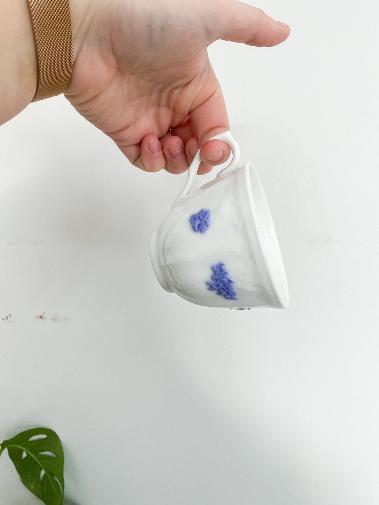 Woman is dangling Adderley tea cup in her hand by the teacups handle. The cup is facing down and shows off its lovely violet flowers in stark constrat to the white teacup. There is a green Monstera leaf in the background