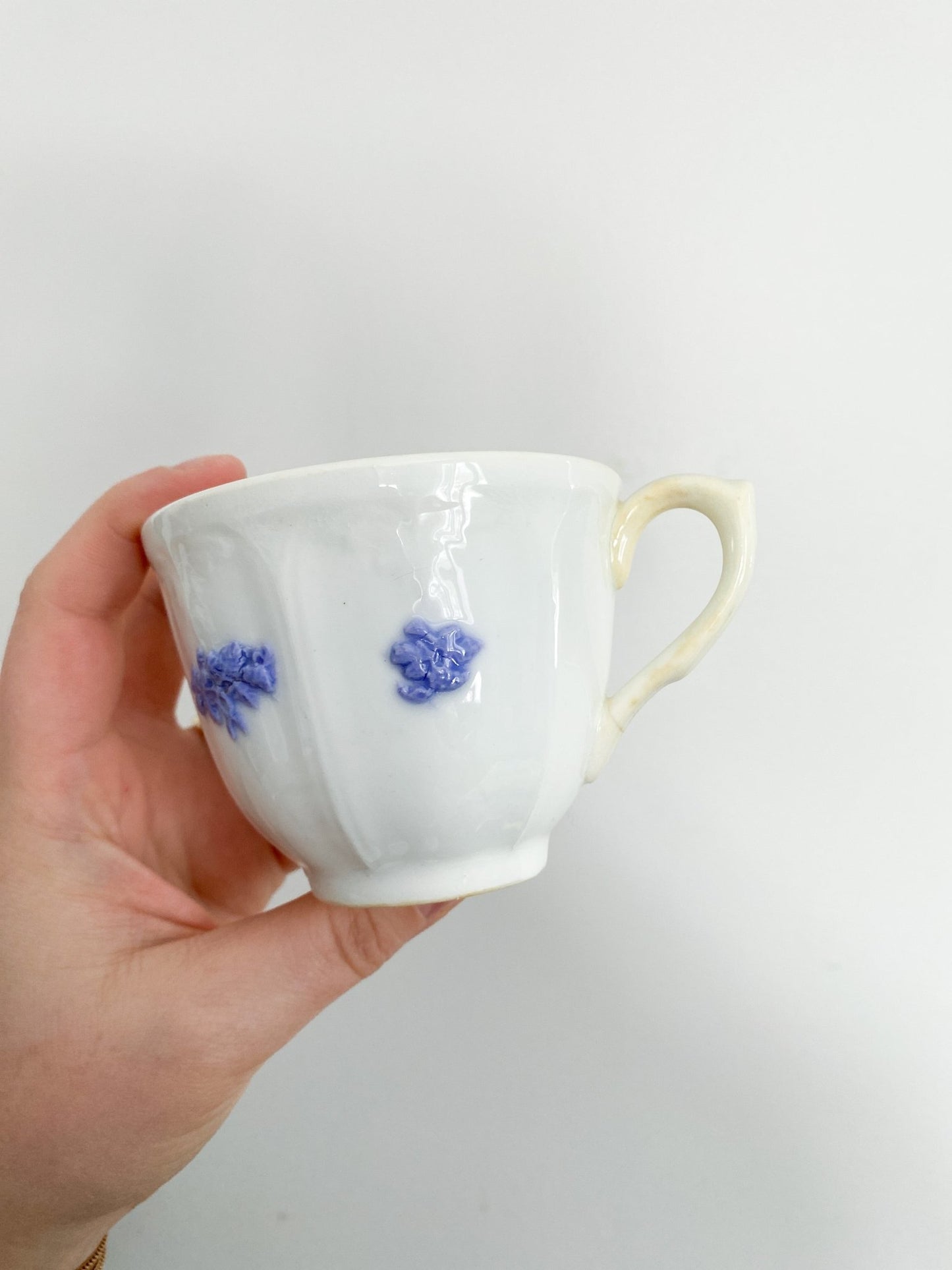 A woman holds the teacup in her hand. The teacup shows off the violet flowers, the handle is slightly yellowing and pointed to the right.