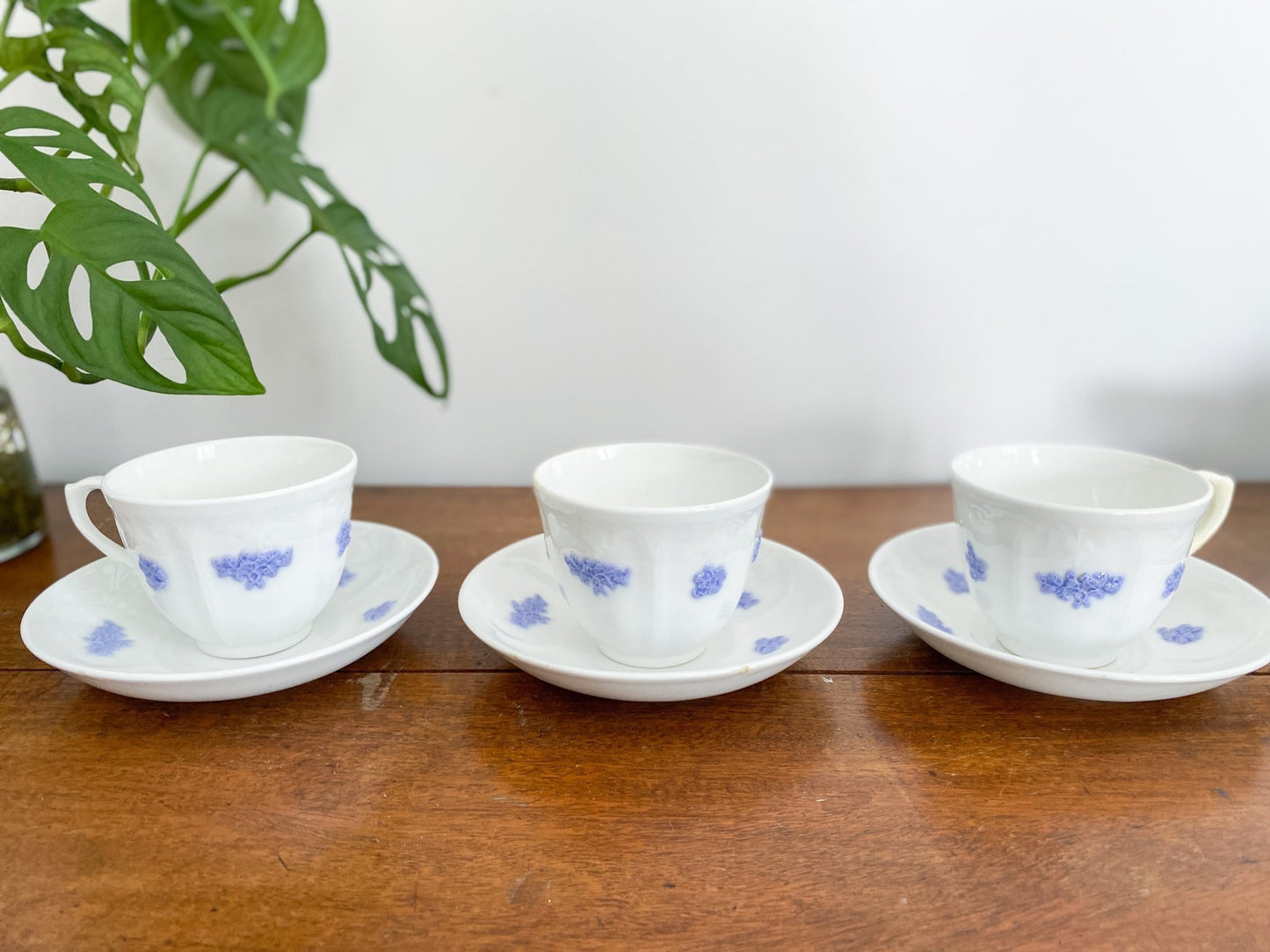 There are three cups in a row on a wooden table. The teacups are all upright but the handle is facing separate directions. The teacups are on the saucers and show the blue or violet colours. The left shows a Monstera plant