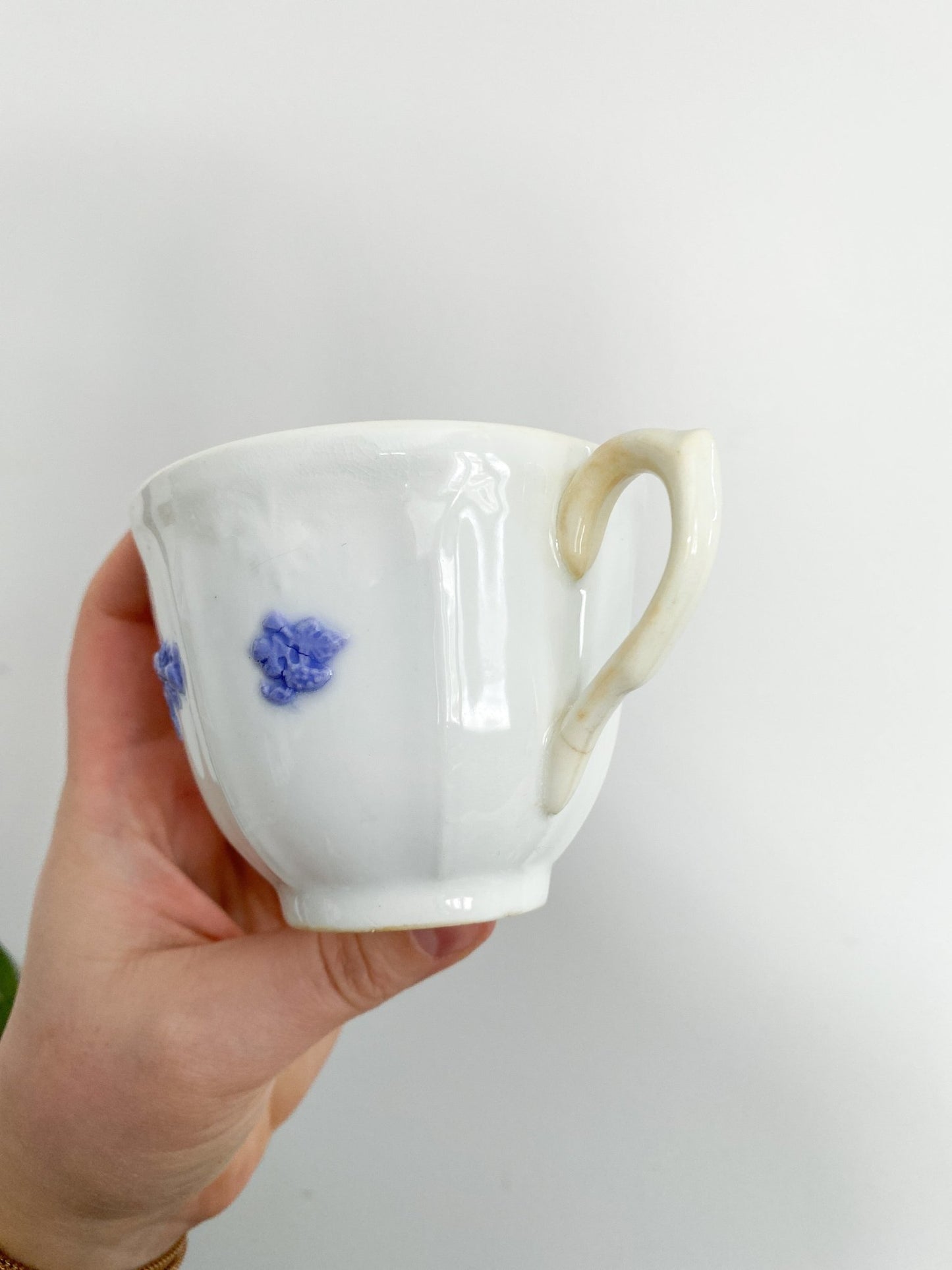 A woman holds the teacup in her hand. The teacup shows off the violet flowers, the handle is slightly yellowing and pointed to the right.