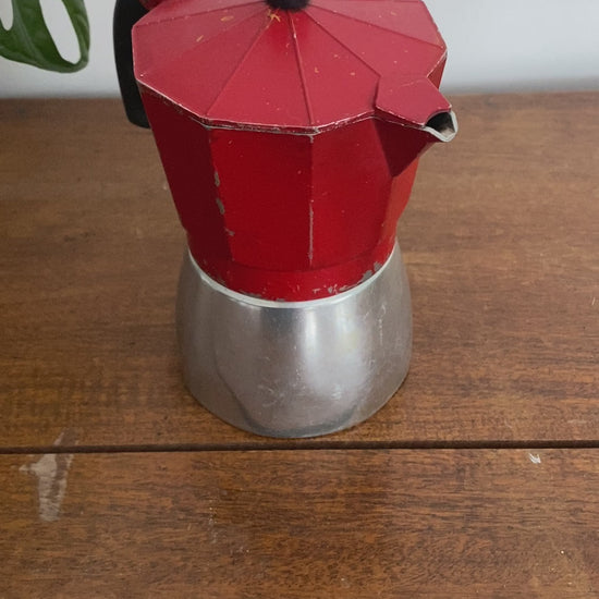 A closeup for the moka pot which is red on the top and silver on the base. The woman removes the top which is on hinges to show the inside. 