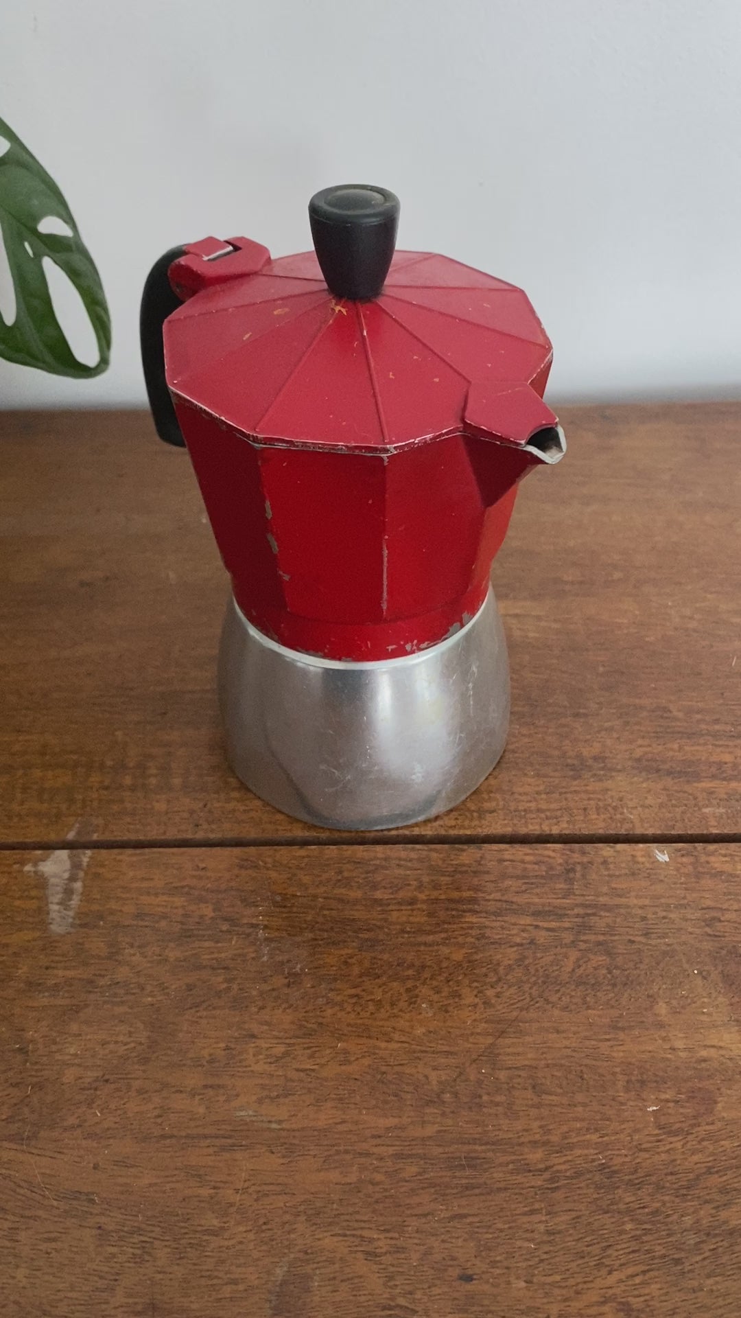 A closeup for the moka pot which is red on the top and silver on the base. The woman removes the top which is on hinges to show the inside. 