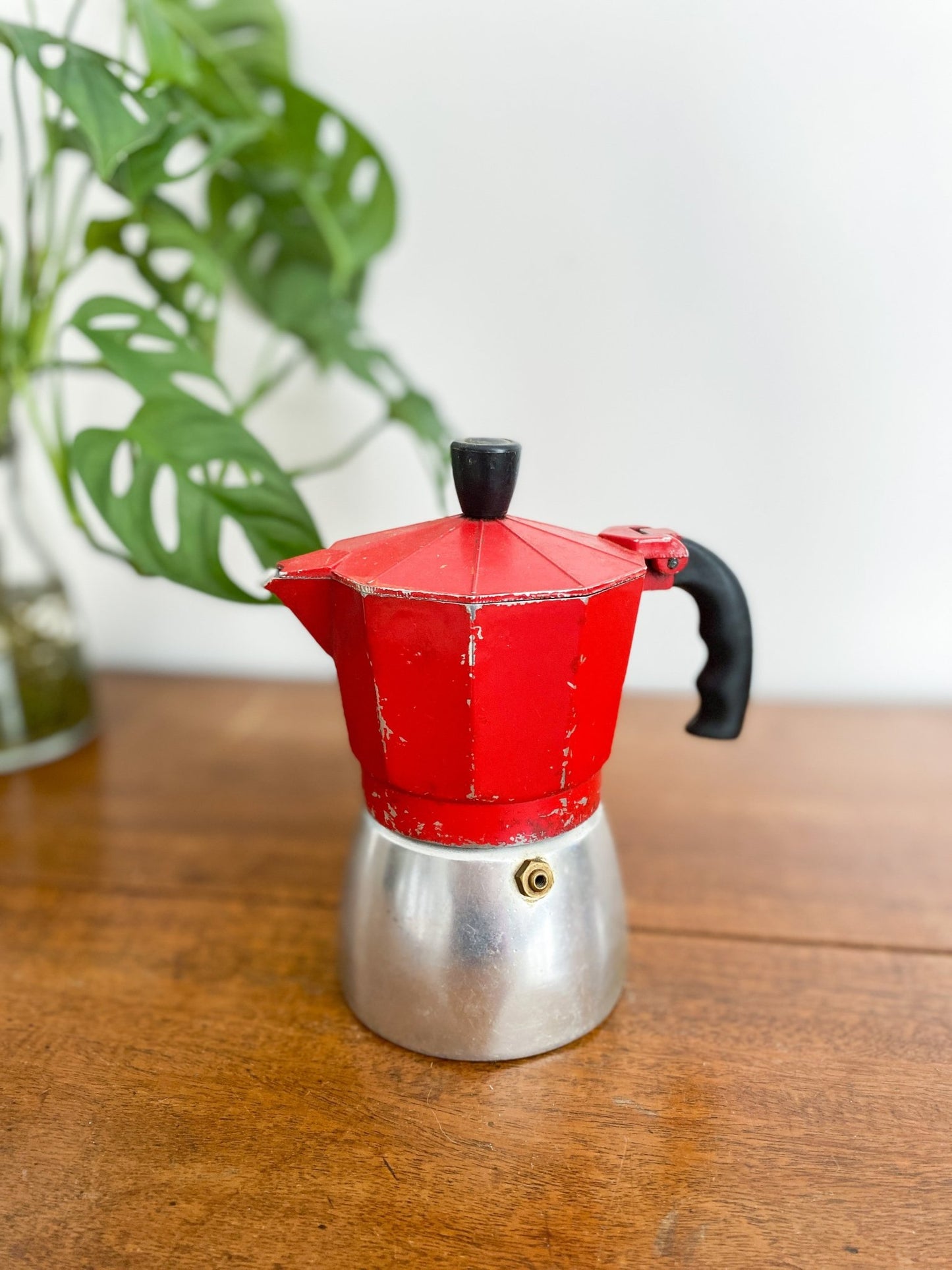 A closeup of the red moka pot, showing some age with the red paint chipping off. The handle is all black and the base is a silver colour. There is a monstera plant to the left.