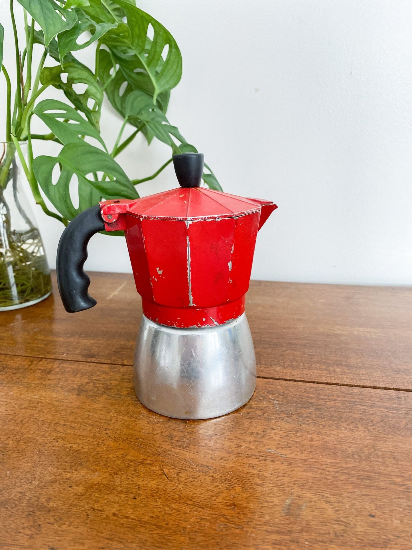 A closeup of the red moka pot, showing some age with the red paint chipping off. The handle is all black and the base is a silver colour. There is a monstera plant to the left.