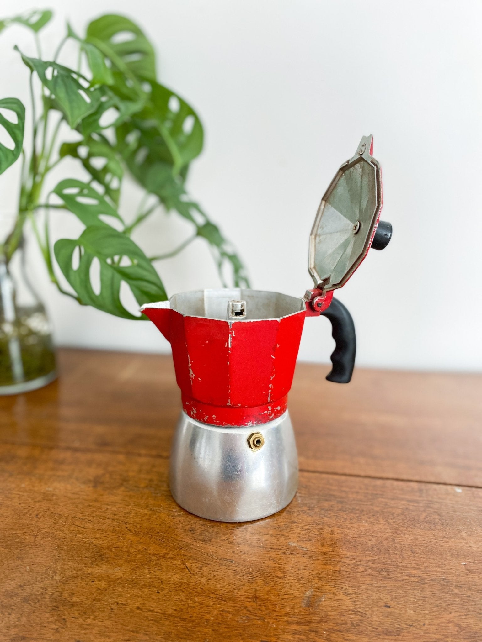 A closeup of the red moka pot facing left, showing some age with the red paint chipping off. The lid of the moka pot is open to show the inside. The handle is all black and the base is a silver colour. There is a monstera plant to the left.