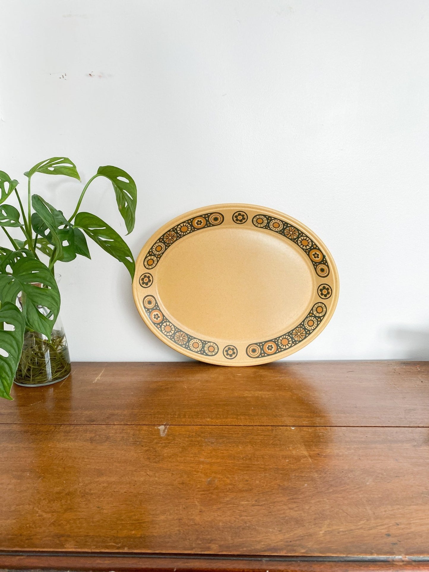 The serving tray is faraway to show the size. The serving tray is different colours of brown, honey and burnt orange with some black flowers rimmed around the edge. A monstera plant is to the left.