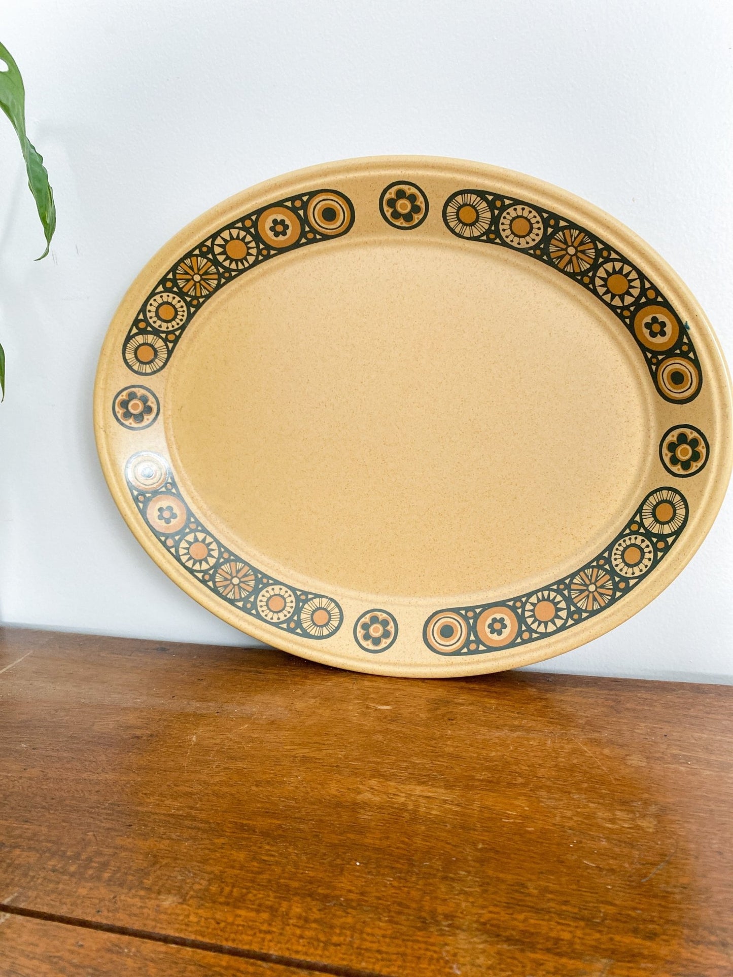 The serving tray is upright against the wall on the wooden table. The tray is a ceramic in a muted honey colour, with flowers along the rim outlined in black and filled in with burnt orange. 