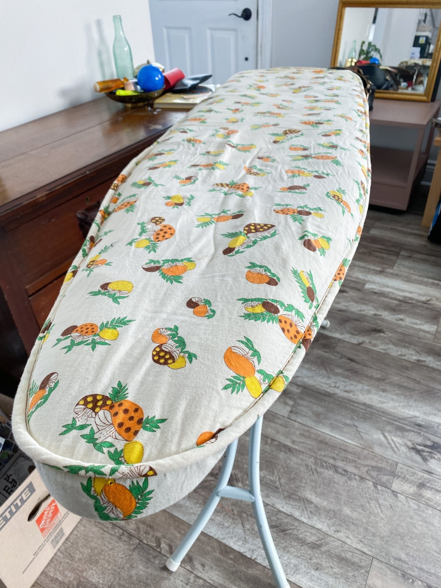 The mushroom fabric on the ironing board, noted are the edges that are expertly sewn. The cover fits over a standard ironing board and the fabric includes colours of orange, brown and yellow mushrooms. 
