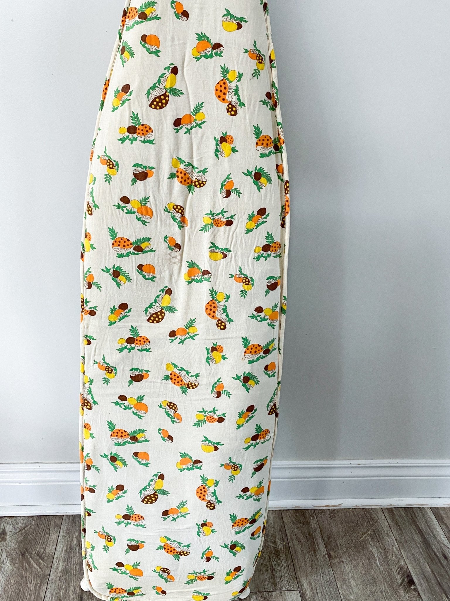 Nearly full frontal of the ironing board cover perfectly fitting over a standard sized ironing board. The pattern is busy with yellow, orange and brown mushrooms of various but similar sizes.