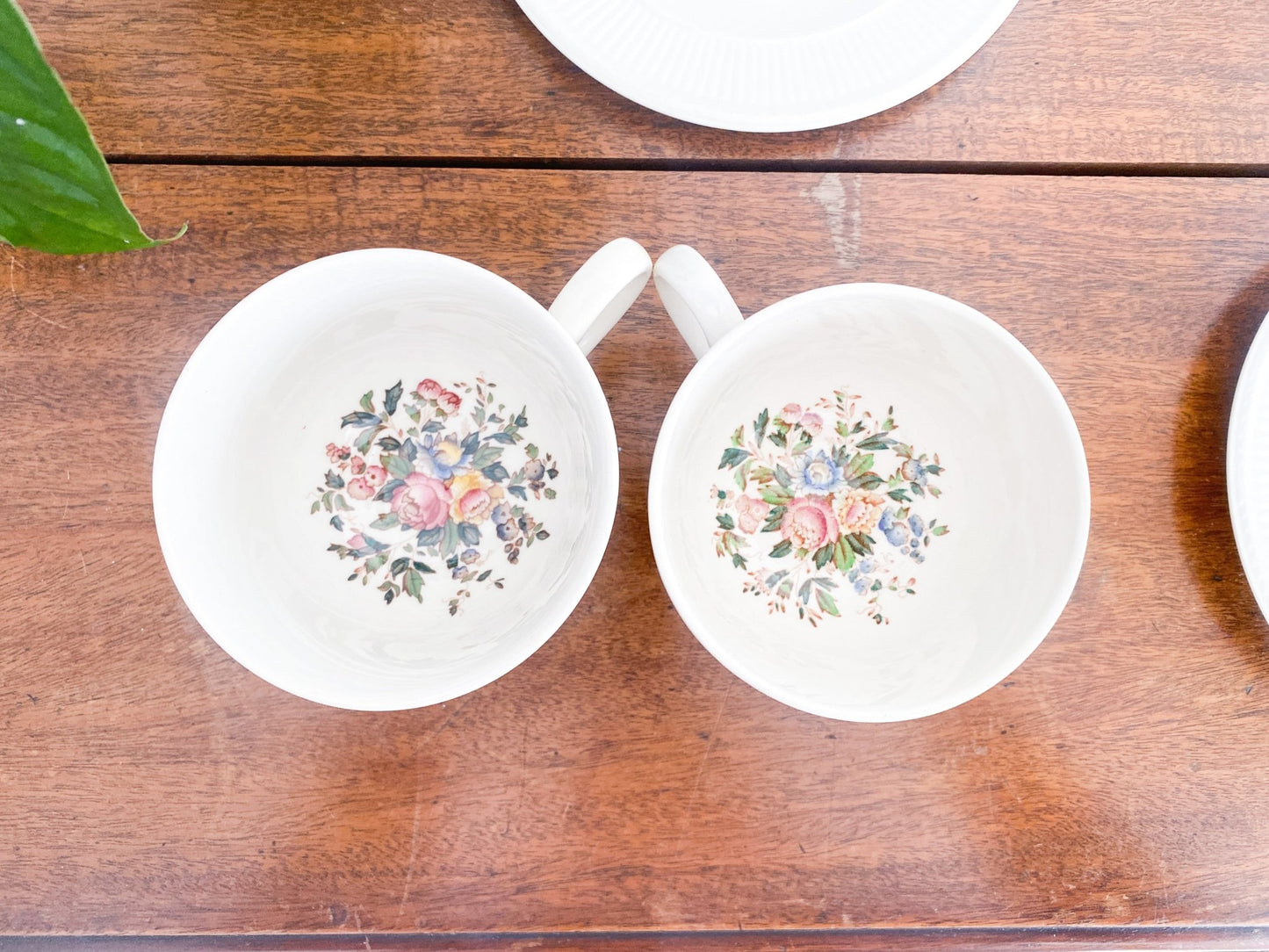 Two cups are touching, both standing upright with the camera to show the interior. The top shows a small portion of a lunch plate and to the right there is a small portion of the saucer. The top left shows a leaf. 