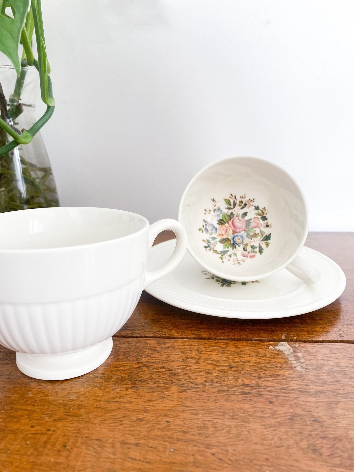 The teacup sits on top of the lunch plate on its side which shows the floral pattern in the centre. There is a teacup standing upright to the left and a monstera plant right above that. 