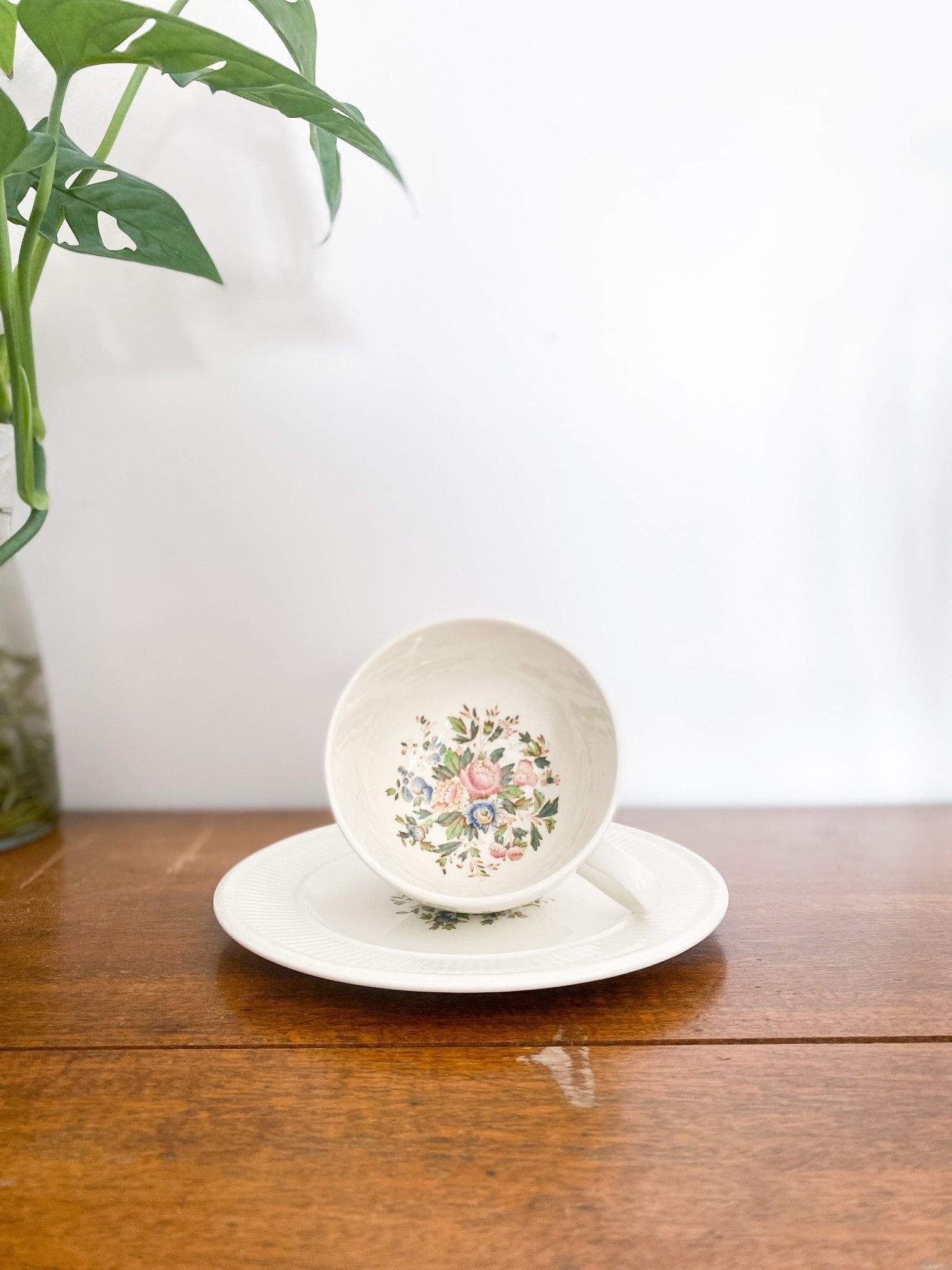 A teacup sits on top of a lunch plate, it shows the interior of the cup with a spread of flowers on the bottom. The left shows a Monstera plate. 